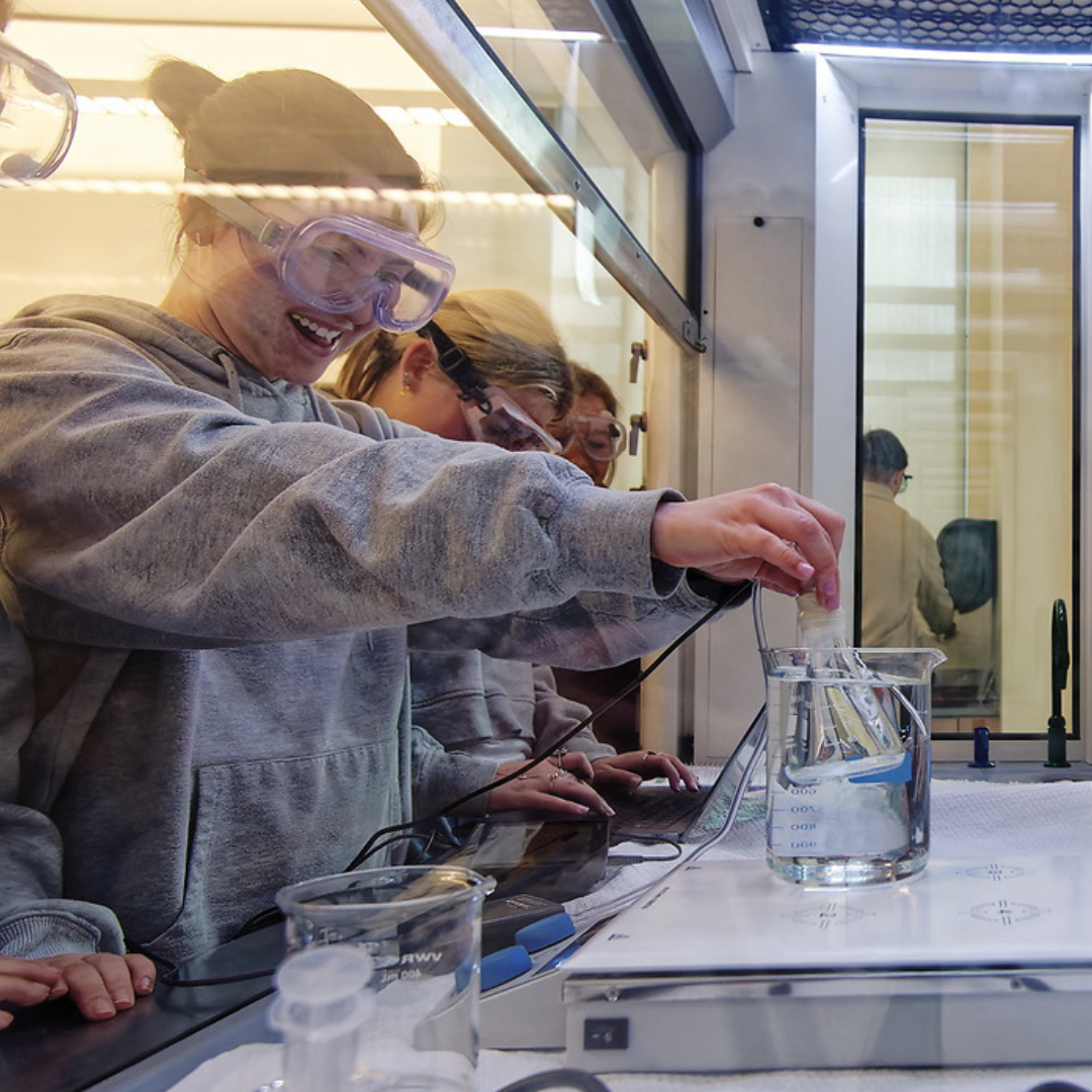 A student in lab gear submerges a beaker into a cylinder of liquid