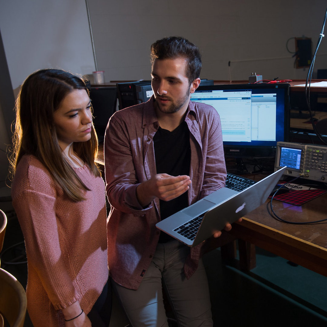 Two telecommunications interns discuss a topic in a dimly lit computer lab