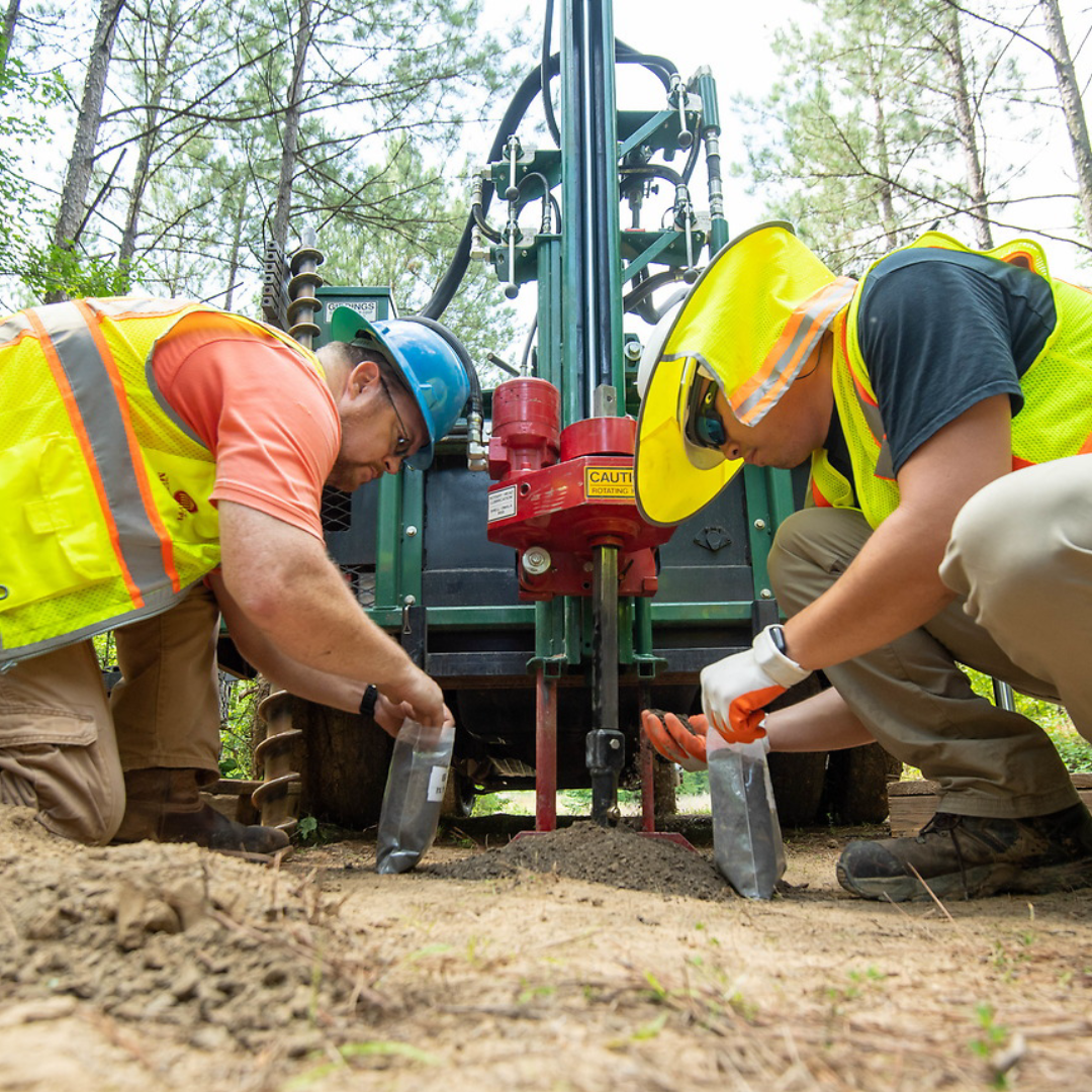 Two researchers from MMRI in hi-vis and hard hats work with a large drilling machine outdoors