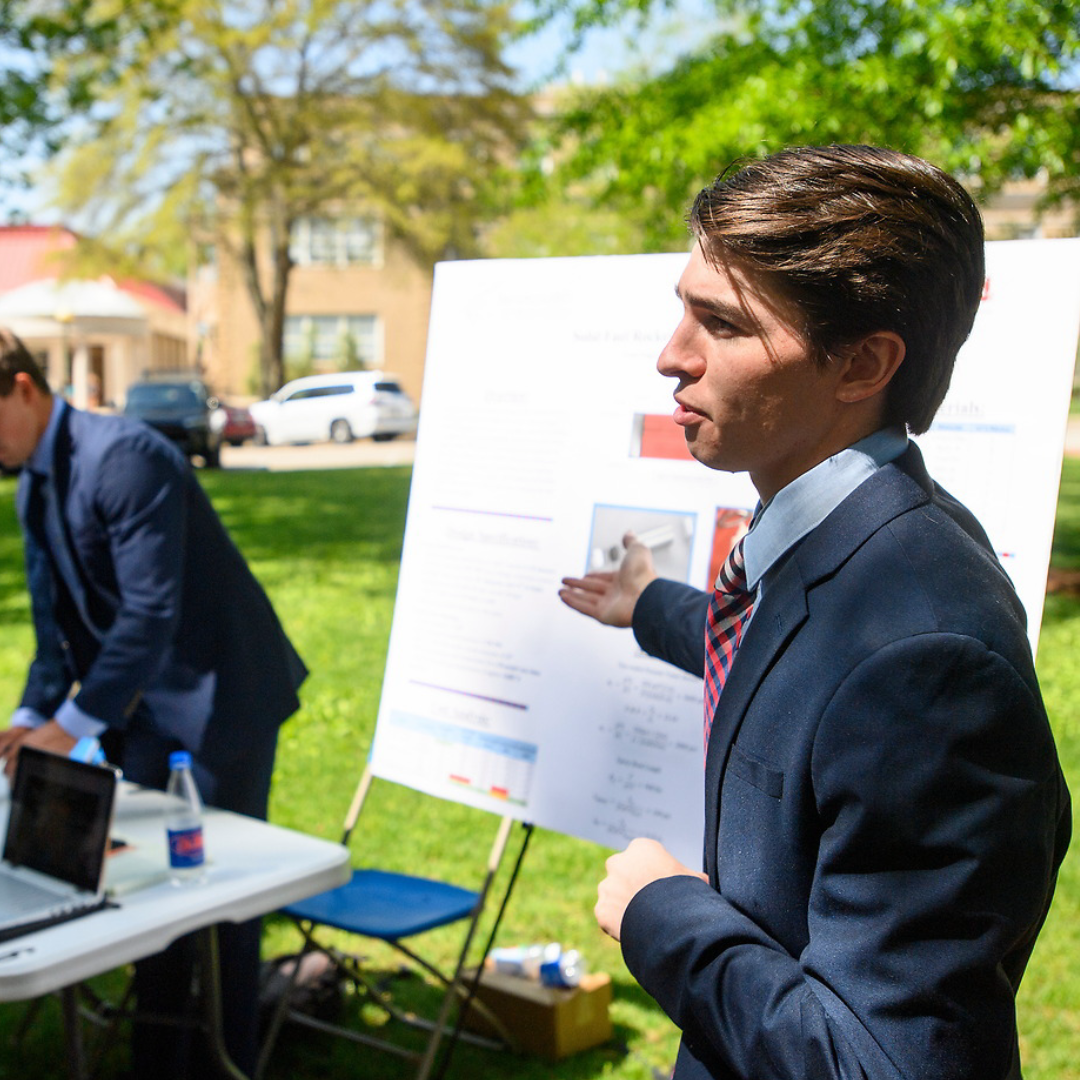 A student dressed in professional business attire presents his project outdoors on the circle in front of Brevard Hall on Ole Miss's campus