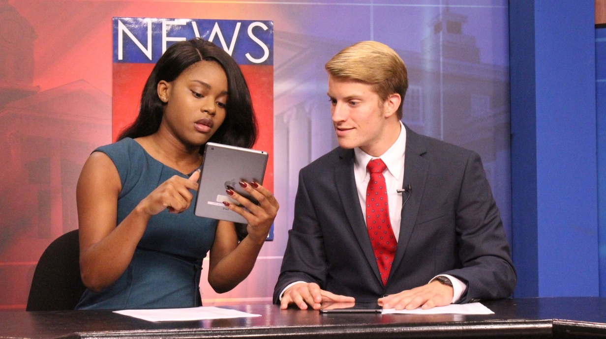 Two students look at a tablet while sitting at the NewsWatch television anchor desk.