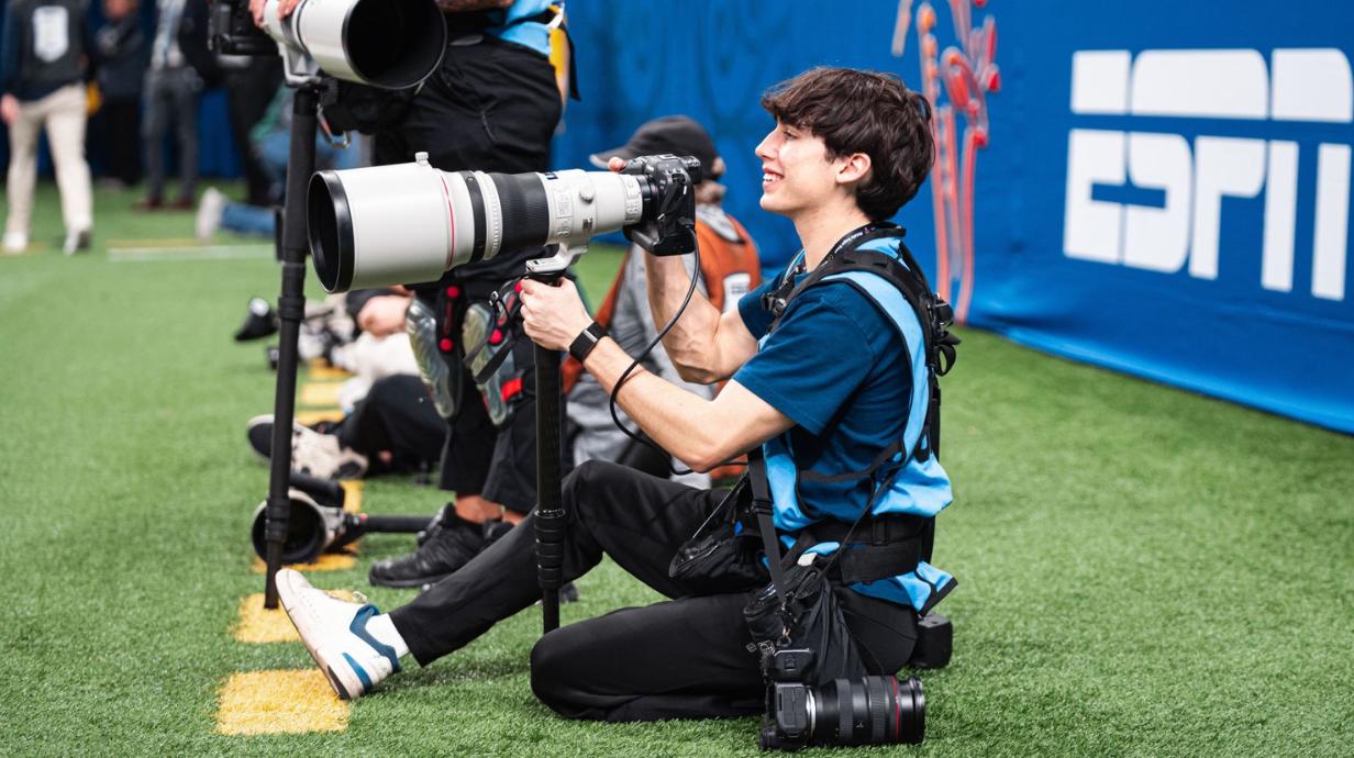 Student sits on football sideline smiling with camera in hand