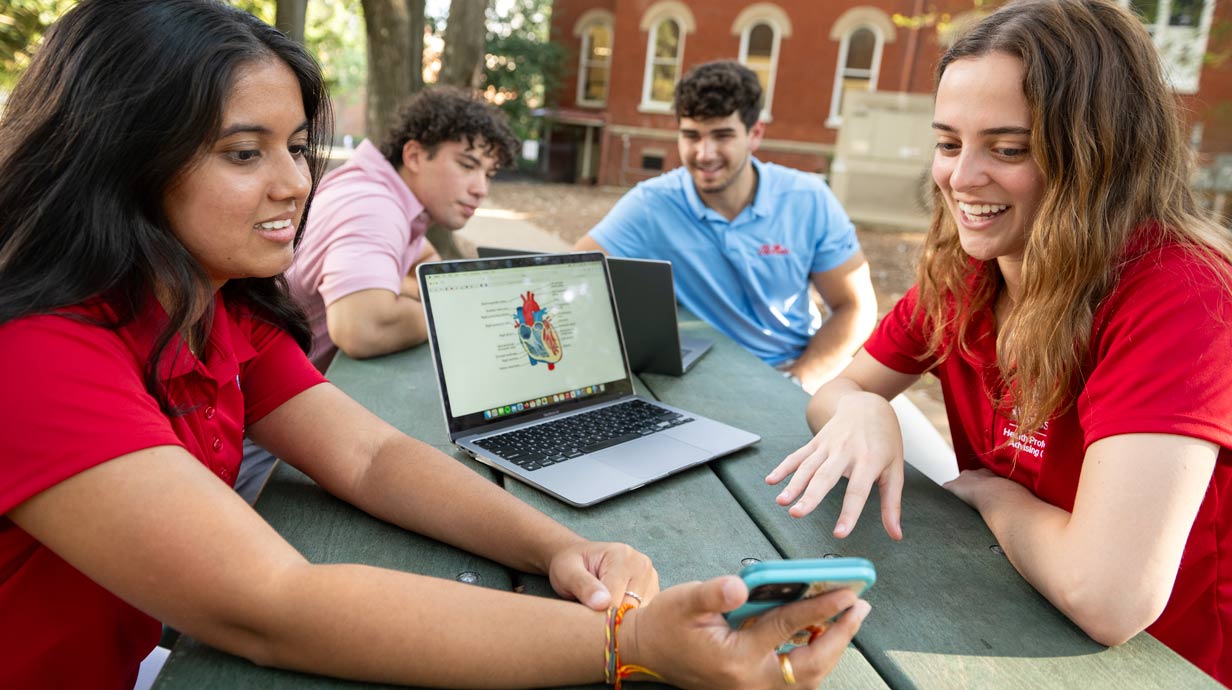 Four Health Professions Advising Office students sitting at a picnic table in the grove looking at a computer and a mobile phone.