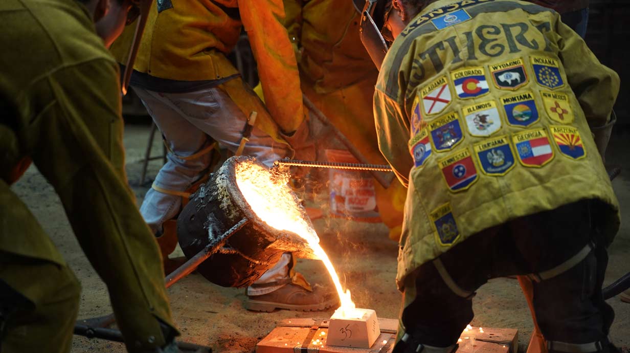 Students participating in an iron pour. Students are wearing bright yellow protective coats and pants as well as helmets with face shields. Bright yellow-orange molten metal is being poured into a mold.