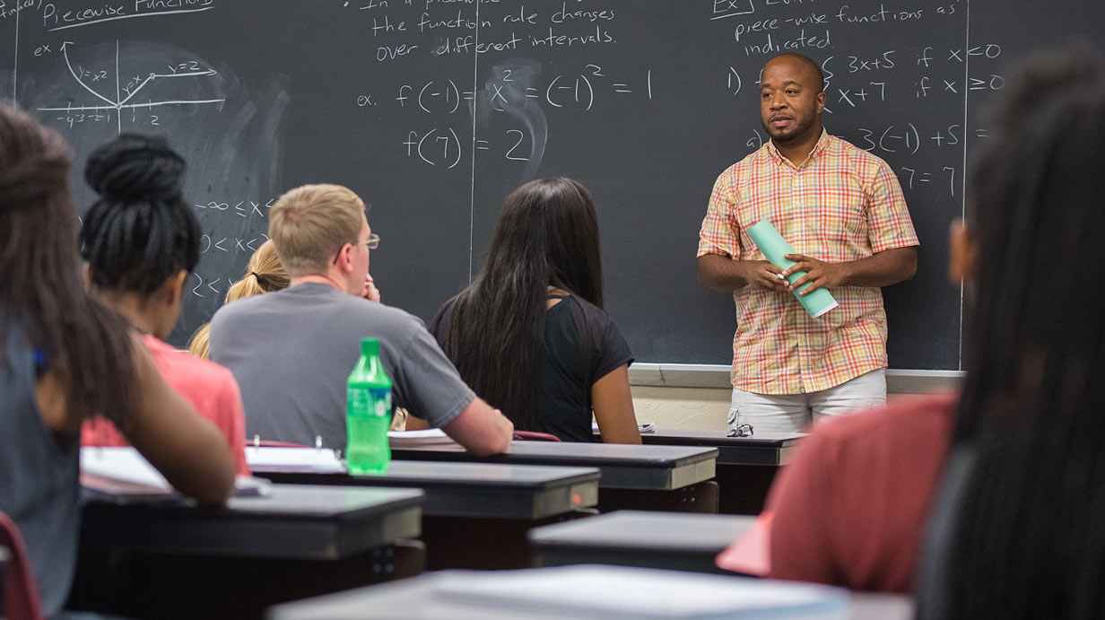 Professor standing in front of chalkboard with numerical expressions written on it; class in foreground.