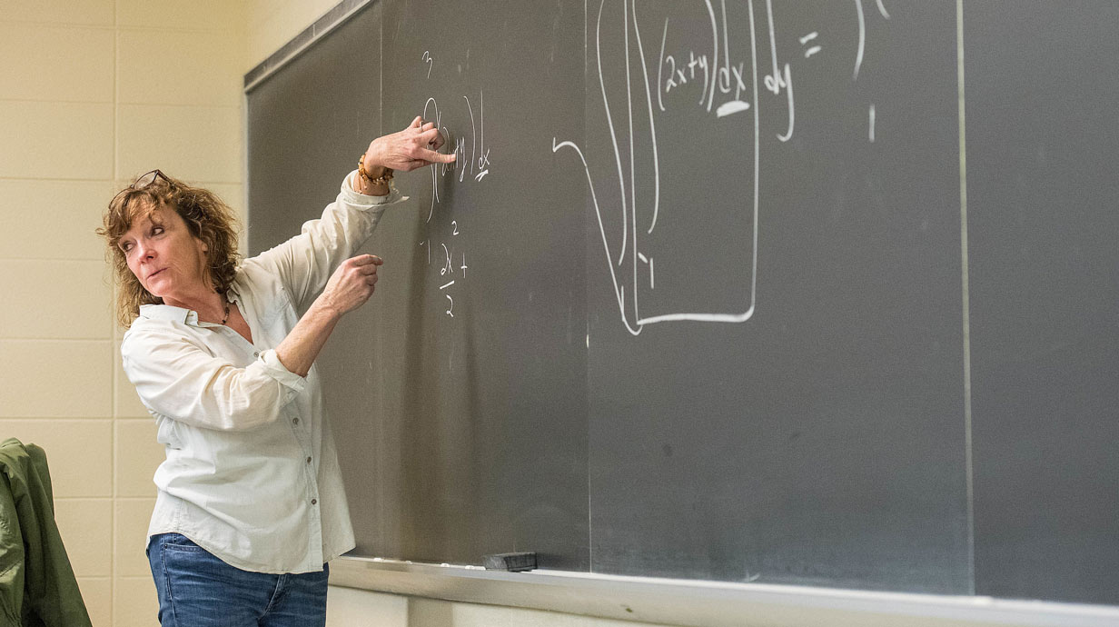 Professor at chalkboard gesticulating with hands and turning to class.