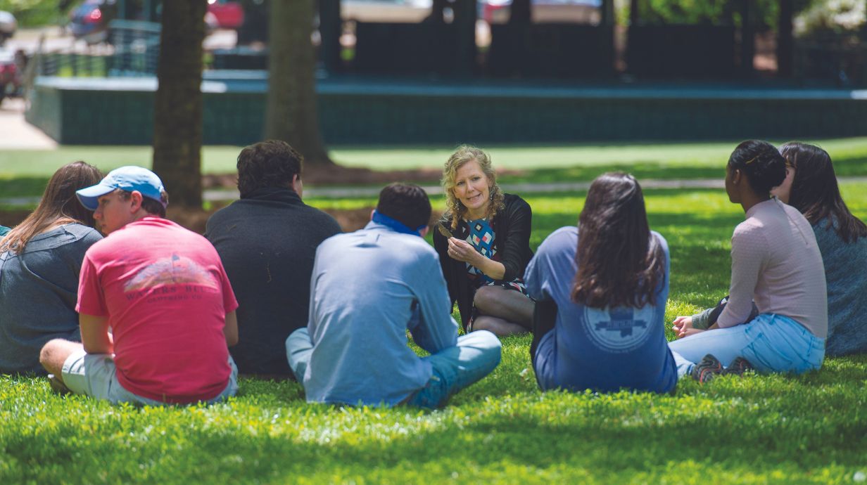 Students listen to a teacher lecture while sitting in the grove. 
