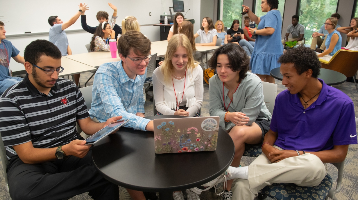 Diverse group of students collaborating around a laptop with floral design, one student using a tablet.