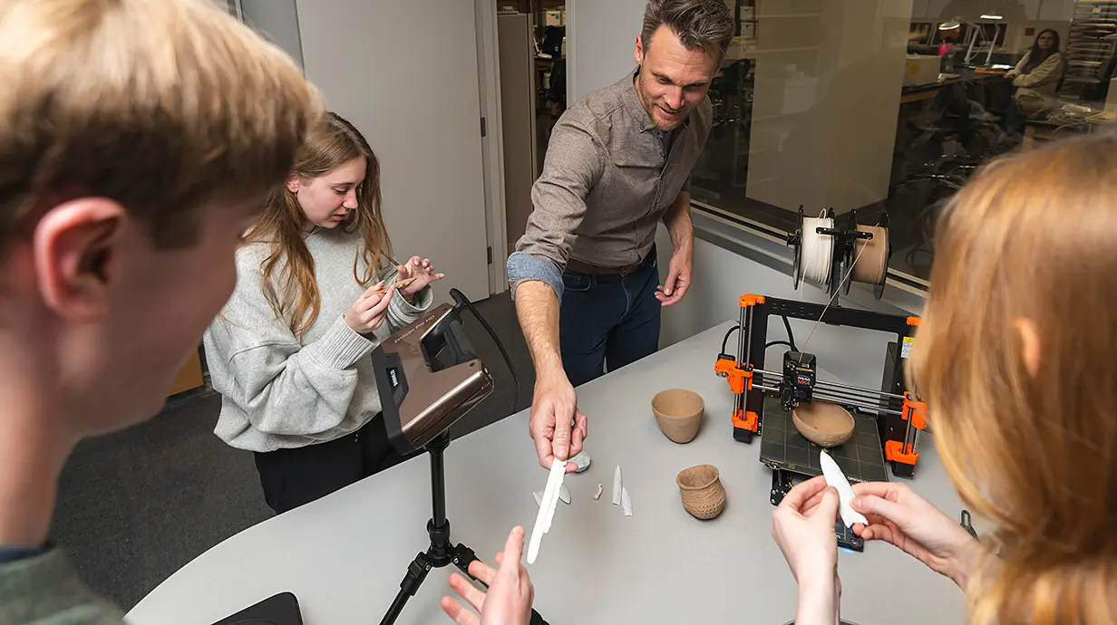 group of people around a table; machinery on table; two people are looking closely at white pointy objects; one person is handing another person across the middle of the image, a white pointy object