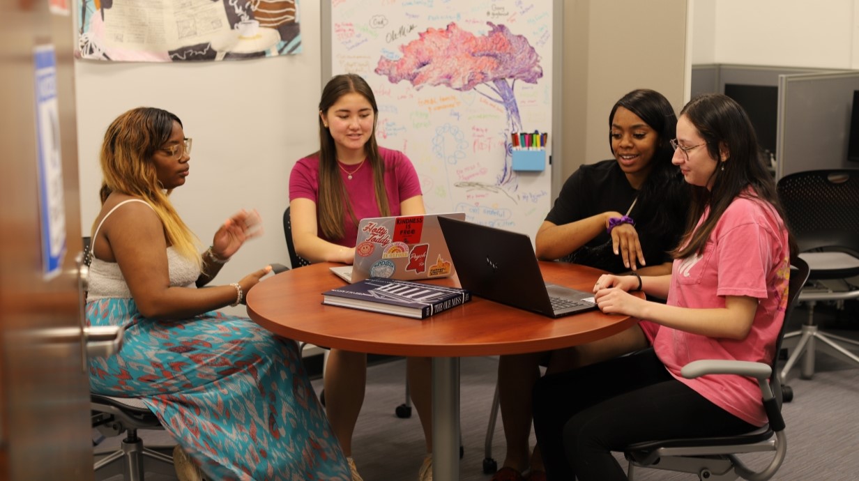 four students are seated together around a table in a psychology lab around a table looking at a laptop computer 