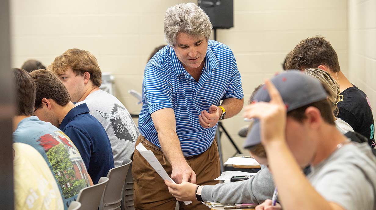 Professor pointing to a piece of paper held by a student. Rows of seated students to the left and right of professor.