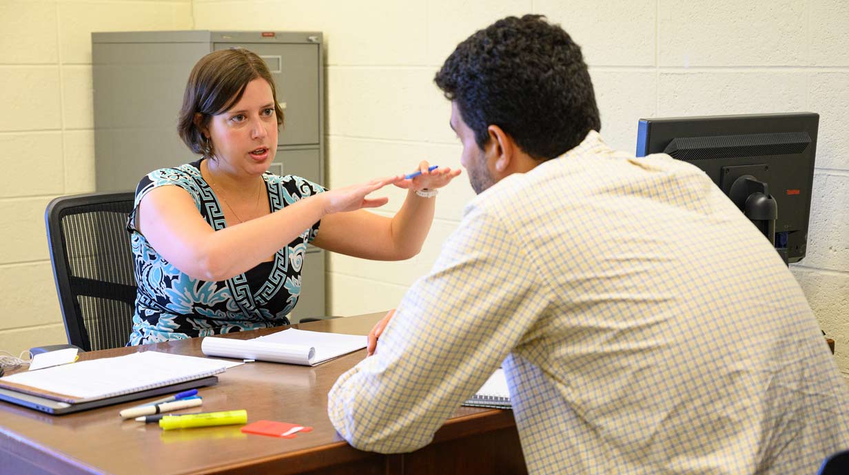 Teacher and student seated on either side of desk. Teacher is gesturing with hands.