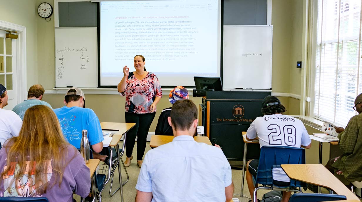 Professor in front of class lecturing. Spanish written on board behind teacher.