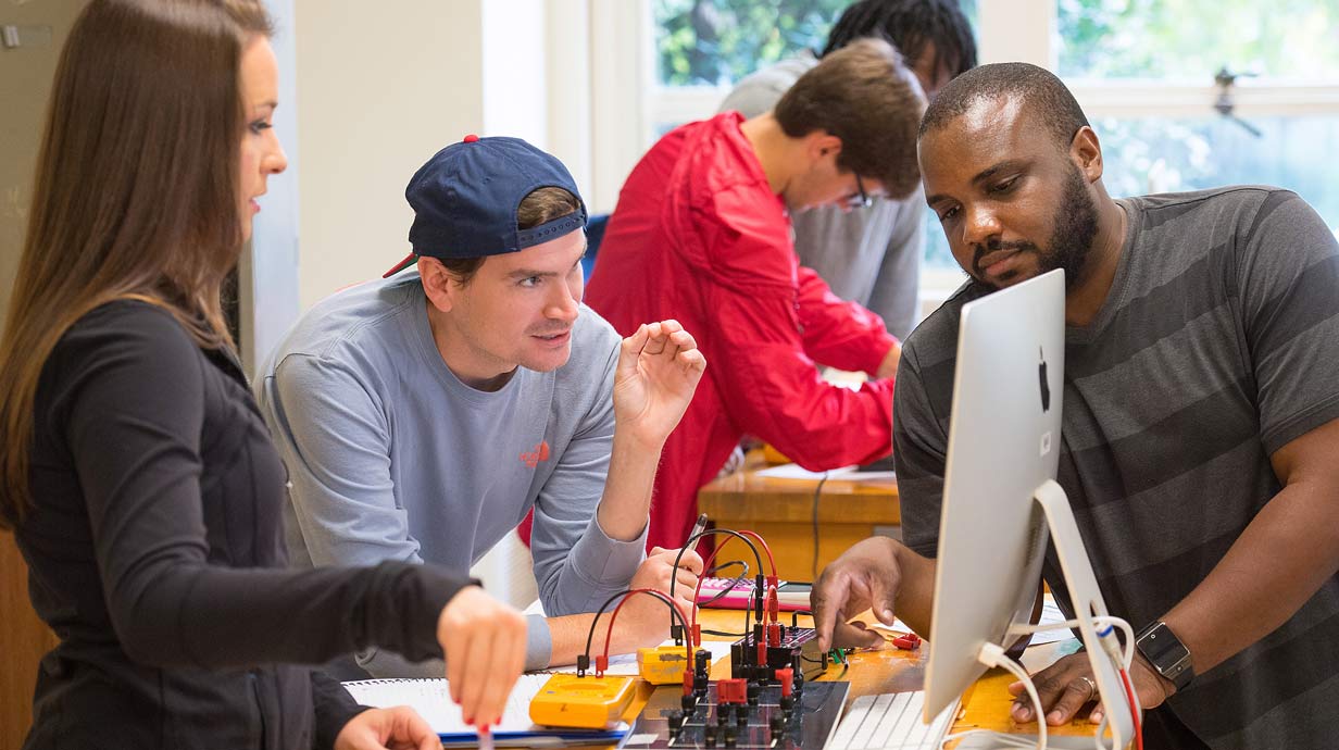 Group of students standing at table looking at monitor. Colorful circuit board model sits on the table.