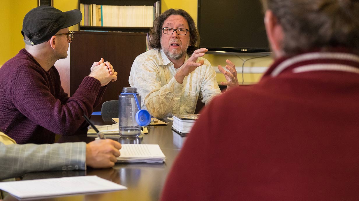 Professor gesticulating with hands at table with two other students.