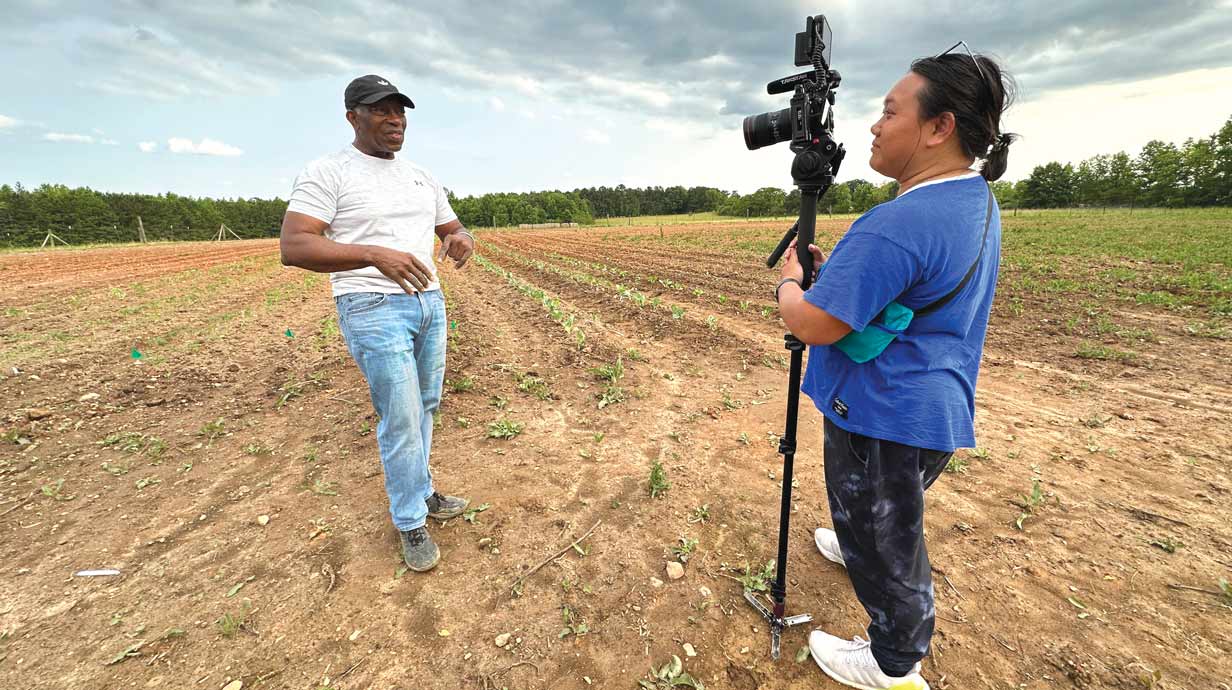 Documentary filmmaker with camera and person being filmed standing in a grassy field.