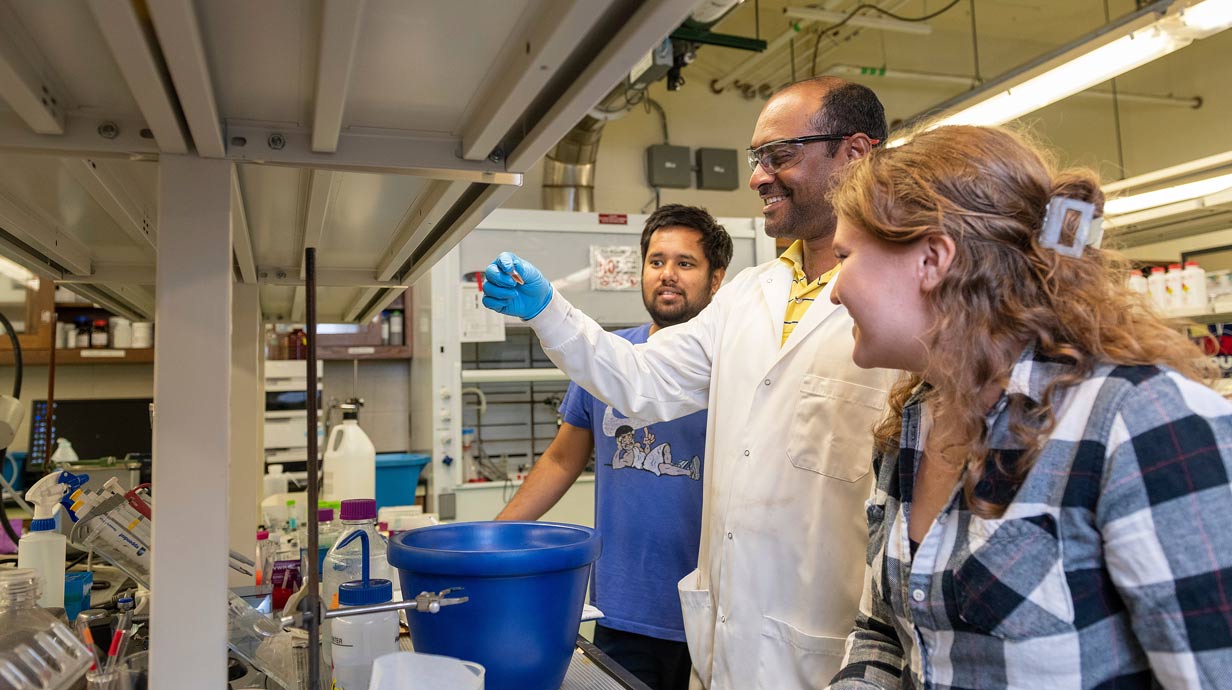 Professor in lab holding up a specimen while with two students on either side of him look on.