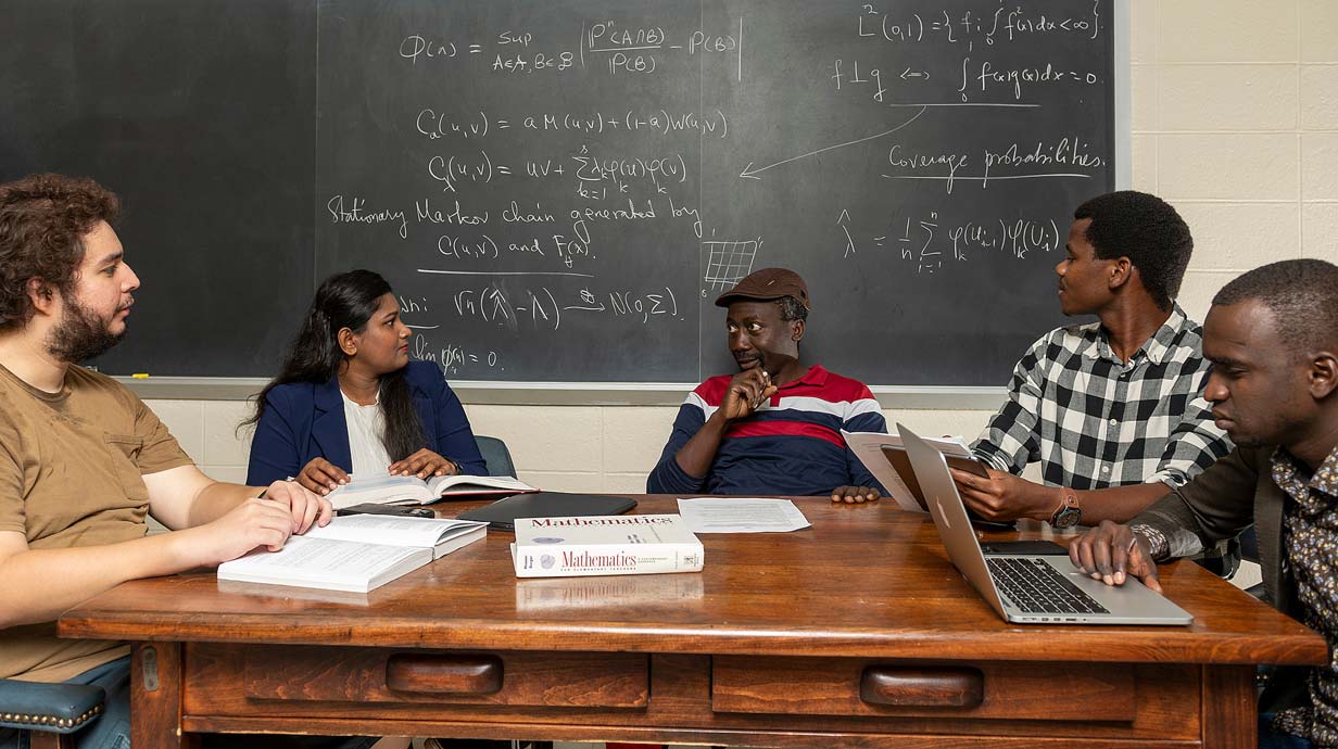 Group of four students and one professor sitting at a table having a discussion. Chalkboard in background is covered with math equations.