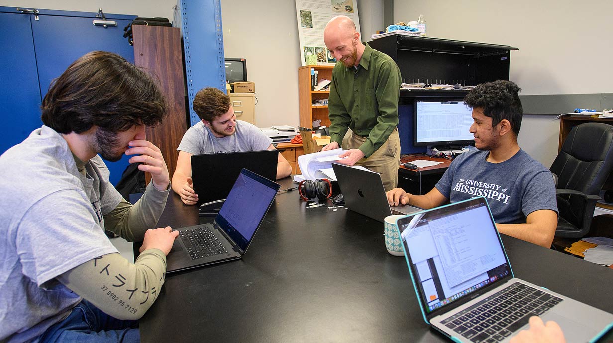 Four students at table are seated with laptop computers. Professor is standing near them thumbing through a book.