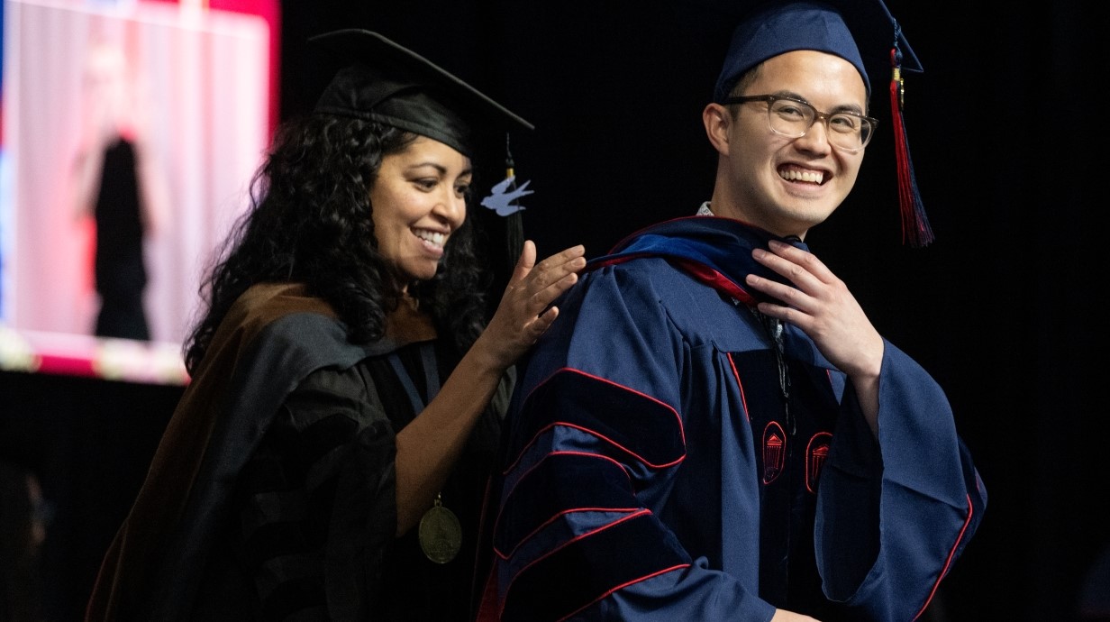 faculty member and student in graduation robes while standing on stage. student is receiving the doctoral hood from a faculty member who is standing behind him.