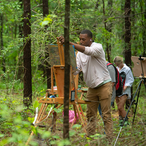 A group of art students paint on easels on-location in the woods.