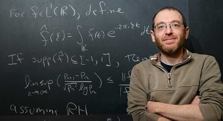 A portrait showing Dr. Micah Milinovich standing next to a blackboard showing some of his recent number theory research.
