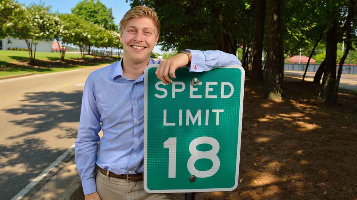 A student poses next to a Manning Speed Limit 18 MPG sign.